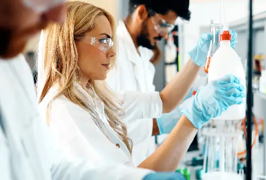 Laboratory technicians in safety glasses examining a specimen bottle, representing pharmacogenomic (PGX) testing of drug metabolism genes.